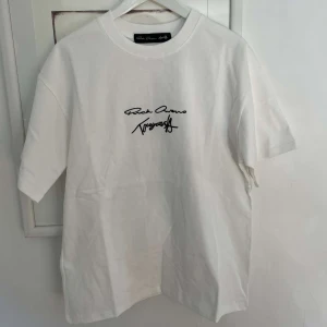 Vit t-shirt med tryck från Rick Owens - Säljer denna snygga vita t-shirt från Rick Owens med svart tryckt text framtill och större tryck på ryggen. Perfekt för en stilren och trendig look.