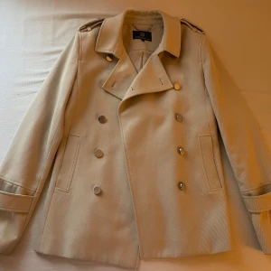 Beige kappa från STOCKH LM - Säljer en stilren beige kappa från STOCKH LM med dubbelknäppning och axelklaffar. Jackan har långa ärmar, klassisk krage och dekorativa knappar med märkets logga. Perfekt för dig som gillar en tidlös look.