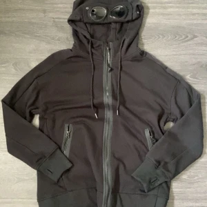 Svart hoodie C.P. Company Medium - Säljer en svart hoodie från C.P. Company med dragkedja framtill och två fickor med dragkedja. Huvan har inbyggda glasögon och snörning. Har inte använts mycket alls, nyskick