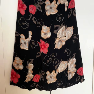 Svart blommig långkjol - Svart långkjol med stora blommor i rött, beige och vitt. Kjolen har en lätt utställd form och ett mjukt, följsamt material. Perfekt för dig som vill ha en färgstark och elegant look.