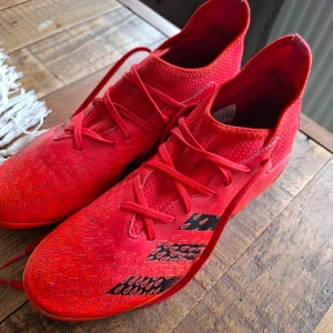 Adidas Predator fotbollsskor i rött - Säljer ett par röda Adidas Predator fotbollsskor med snörning och svarta detaljer. Skorna har en stickad ovandel i syntetmaterial och är designade för bra grepp på planen. Perfekta för dig som vill sticka ut på fotbollsplanen!
