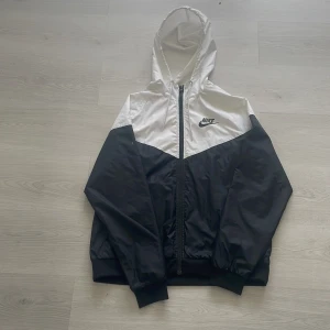 Svart och vit vindjacka från Nike - Säljer en snygg svart och vit vindjacka från Nike med huva och dragkedja. Jackan har elastiska muddar och klassisk Nike-logga på bröstet. Perfekt för dig som gillar sportig stil och vill ha något lätt att slänga på sig.