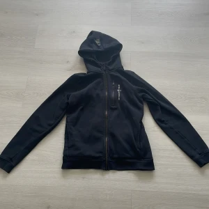 Svart hoodie från Sail Racing - Svart hoodie från Sail Racing med dragkedja framtill och en bröstficka med dragkedja. Märkets logga är broderad på bröstet och på huvan. Perfekt för en sportig och avslappnad stil.