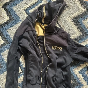 Svart hoodiejacka från BOSS passar L och M - Svart hoodiejacka från BOSS med gulddetaljer och dragkedja. Jackan har huva med snörning och logotyp på bröstet. Perfekt för en avslappnad och sportig stil