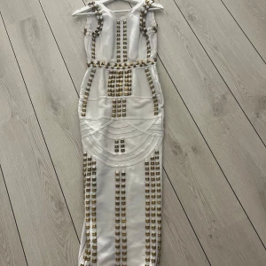 Vit långklänning med gulddetaljer - Säljer en vit långklänning med unika guldfärgade detaljer och geometriska mönster. Klänningen har smala axelband och en figurnära passform som framhäver siluetten. Perfekt för speciella tillfällen när du vill sticka ut.