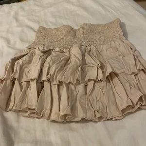 Beige volangkjol från SHEIN - Supersöt beige volangkjol från SHEIN med smock i midjan och flera lager volanger. Kjolen är kort och har en luftig känsla, perfekt för en trendig och avslappnad stil. Passar dig som gillar feminina detaljer och mjuka toner.