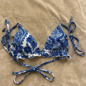 Blåvit bikiniöverdel med paisleymönster - Säljer en snygg bikiniöverdel i blått och vitt med paisleyinspirerat mönster. Modellen har trekantskupor och knytband i både nacke och rygg för justerbar passform. Perfekt för stranden eller poolen!