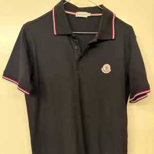 Svart pikétröja från - Snygg svart pikétröja med klassisk krage och knappar. Tröjan har Moncler-logga på bröstet och rödvita detaljer på krage och ärmslut. Perfekt för en stilren look.