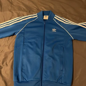 Blå Adidas track jacket med vita ränder - Säljer en klassisk blå Adidas track jacket med vita ränder längs ärmarna och vit Adidas-logga på bröstet. Jackan har dragkedja framtill, ståkrage och två fickor. Perfekt för en sportig och avslappnad stil.