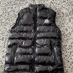 Svart väst från moncler - Snygg svart dunväst från Moncler med glansig finish och klassisk logotyp på bröstet. Västen har hög krage, dragkedja framtill och två fickor med dragkedja. Bra för sommar i Sverige