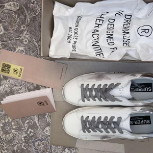 Golden Goose sneakers med silverdetaljer - Säljer ett par vita Golden Goose sneakers med grå snörning, silverfärgade detaljer på hälen och den klassiska stjärnan på sidan. Skorna har en platt sula och rund tå, och är tillverkade i skinn med slitna detaljer för en cool look. Priset är ej hugget i sten!