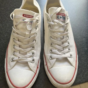 Vita Converse All Star sneakers - Klassiska vita Converse All Star sneakers med lågt skaft och röd rand längs sulan. Skorna har snörning och rund tå, perfekt för en avslappnad och tidlös stil.