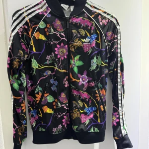 Blommig set från Adidas - Säljer Adidas set med färgglatt blommigt mönster och fjärilar. Jackan har dragkedja framtill, vita ränder på ärmarna och ribbade muddar. Perfekt för dig som vill sticka ut med en unik och sportig stil.