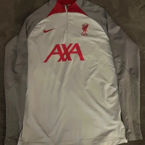 Liverpool träningströja från Nike - Säljer en långärmad träningströja från Nike med Liverpool FC-logga och AXA-tryck på bröstet. Tröjan har halv dragkedja och är i slim fit-modell, perfekt för fotbollsträning. Finns inga synliga defekter.