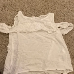 Vit offshoulder topp från H&M - Säljer en söt vit offshoulder topp från H&M i storlek 140. Toppen har volangdetalj längs axlarna och är ärmlös, perfekt för varma dagar. Enkel och stilren design som passar till mycket.