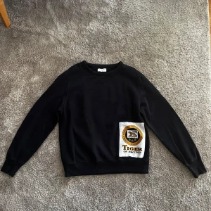 Svart sweatshirt från Tiger of Sweden - Svart sweatshirt från Tiger of Sweden med rund halsringning och lång ärm. Tröjan har en stor vit patch med logga och texten 'Tiger of Sweden' framtill. Perfekt för en stilren och avslappnad look.
