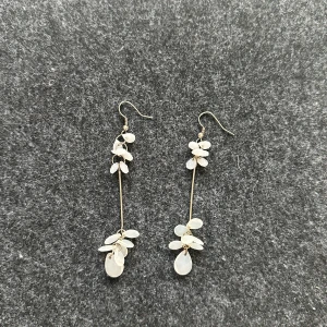 Långa örhängen med vita pärldetaljer - Säljer ett par eleganta, långa örhängen med tunna guldfärgade stänger och vita pärlliknande stenar i blomform. Perfekta för att ge en stilren touch till din look. Krokar i metall för enkel användning.