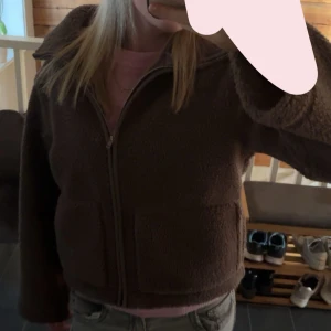 Brun teddyjacka med dragkedja i storleken S - Mysig brun teddyjacka med krage och dragkedja framtill. Jackan har lång ärm och en lite boxig passform som känns både trendig och bekväm. Perfekt för kyligare dagar när du vill ha något mjukt och stilrent.