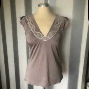 topp med spetsdetalj S viskos - Söt beige topp med v-ringning och dekorativ spets längs urringningen. Toppen har korta volangärmar och är tillverkad i ett mjukt, lätt material. Perfekt för en feminin och stilren look.