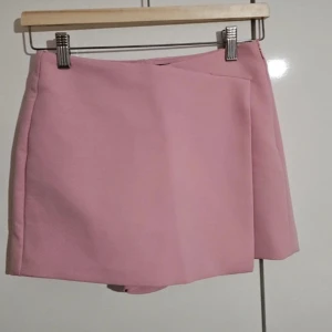 Rosa omlottkjol med shorts - Säljer en ljusrosa omlottkjol med shorts under, perfekt för dig som vill ha kjol-looken men ändå känna dig bekväm. Kjolen har en stilren design utan synliga detaljer framtill och två fuskfickor bak.