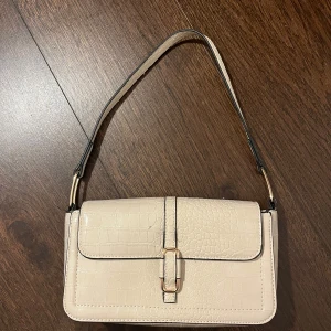 Beige axelväska med krokodilpräglat mönster - Säljer en snygg beige axelväska med krokodilpräglat mönster och guldfärgat spänne framtill. 