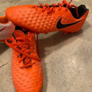 Orangea Nike fotbollsskor med svarta detaljer - Säljer ett par orangea Nike fotbollsskor med svarta detaljer och snörning. Skorna har en prickig textur på ovandelen och en låg profil, perfekt för spel på gräs. Klassisk Nike-logga på sidan.