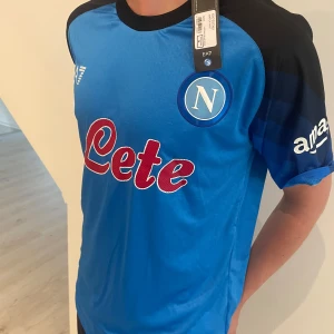 Napoli fotbollströja 2023/24 EA7 - Säljer en officiell Napoli hemmatröja från säsongen 2023/24. Tröjan är blå med svarta axlar och detaljer, har klubbmärket på bröstet och sponsorloggor. Tillverkad i lätt och ventilerande material, perfekt för fotboll eller supporterliv.