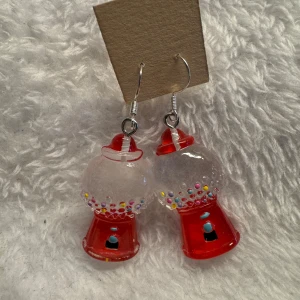 Tuggummiautomat örhängen - Unika örhängen formade som små tuggummiautomater i rött och genomskinligt med färgglada detaljer. Perfekt för dig som vill sticka ut och addera en lekfull touch till din stil. Krokarna är i sterling silver 