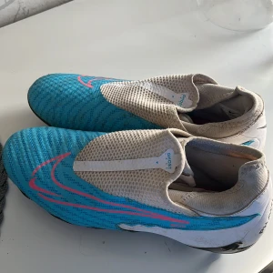 Nike fotbollsskor blå och vita - Säljer ett par blå och vita Nike fotbollsskor med rosa detaljer. Skorna har en strumpliknande passform utan  och är designade för spel på gräs med fasta dobbar undertill. Perfekta för dig som vill sticka ut på planen!äkta 