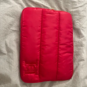 Röd datorväska från Carolina Herrera - Säljer en snygg röd datorväska från Carolina Herrera med broderad logga på framsidan och guldfärgad dragkedja. Väskan har ett vadderat material som skyddar din laptop och en elegant design med märkets signaturdetaljer.