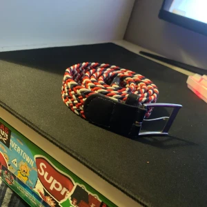 Färgglatt flätat bälte med svart spänne - Säljer ett snyggt flätat bälte i rött, vitt och blått med ett svart metallspänne. Bältet har en sportig och ungdomlig stil och passar perfekt till jeans eller shorts.