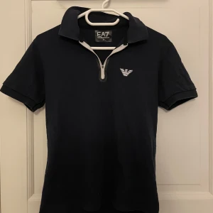 Mörkblå t-shirt med dragkedja från EA7 Emporio Armani - Snygg mörkblå t-shirt från EA7 Emporio Armani med kort dragkedja vid halsen och vit logga på bröstet. Klassisk krage och korta ärmar. Perfekt för en stilren och sportig look.