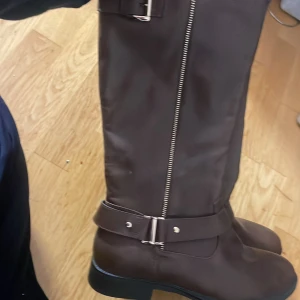 Bruna höga stövlar med dragkedja och bälte - Säljer ett par bruna höga stövlar med dekorativt bälte och dragkedja på sidan. Snygg och stilren design med platt sula, perfekt till många outfits. Storlek 41. Aldrig andvända 🤎🤎