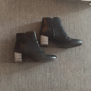 Svarta boots med silvrig klack - Säljer ett par svarta boots med rund tå och en unik silvrig klack med mönstrad struktur. Skorna har dragkedja på sidan och är i ett stilrent utförande som passar till många outfits.