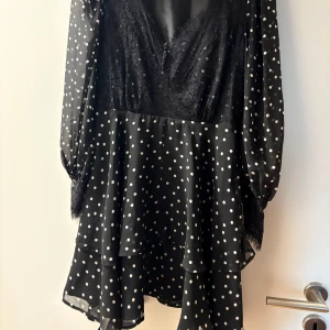 Svart prickig omlottklänning med spets - Supersöt svart klänning med vita prickar och v-ringning. Klänningen har långa, transparenta ärmar och spetsdetaljen. Kjolen är volangig och ger en flowy känsla. Klänningen har en dragkedja på sidan. Den kommer med ett band, ena hålet för bandet har gått av men kan syns tillbaka.