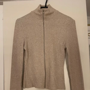 Beige ribbad kofta med dragkedja - Säljer en beige ribbad kofta med dragkedja framtill och hög krage. Tröjan har lång ärm och är i ett mjukt stickat material. Perfekt för lager-på-lager och enkel att matcha med olika outfits.