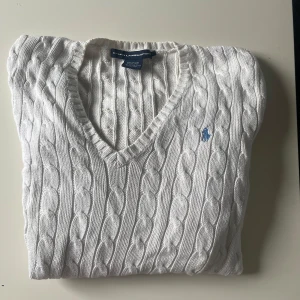 Vit kabelstickad tröja från Ralph Lauren - Säljer en klassisk vit kabelstickad tröja från Ralph Lauren Sport med v-ringning och broderad blå logga på bröstet. Tröjan har lång ärm och är tillverkad i mjuk bomull. Perfekt för en stilren och tidlös look.