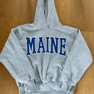 Grå hoodie med MAINE-tryck - Säljer en grå hoodie med blått MAINE-tryck på bröstet. Klassisk modell med huva och ribbade muddar vid ärmslut och nederkant. Perfekt för en avslappnad stil.
