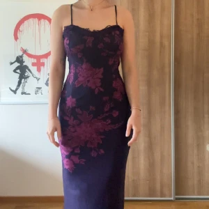 Långklänning med lila blommor Urban Outfitters - Säljer en elegant långklänning med smala axelband och blommigt mönster i lila och mörklila nyanser. Klänningen har figurnära passform och en romantisk känsla, perfekt för speciella tillfällen.