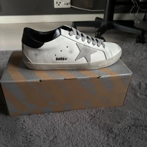Vita sneakers från Golden Goose - Säljer ett par vita sneakers från Golden Goose med silverfärgad stjärna på sidan och grå snörning. Skorna har svart hälparti och klassisk platt sula. Perfekta för dig som gillar en stilren men ändå unik look. Helt nya allt og medföljer. Pris kan diskuteras vid snabb affär‼️