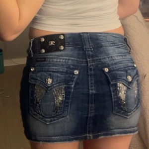 Miss me kjol! - Säljer en blå jeanskjol från Miss Me med glittriga detaljer och dekorativa bakfickor. Kjolen har kort längd, klassisk femficksdesign och silverfärgade knappar och nitar. Perfekt för dig som vill ha en snygg och trendig denimlook.