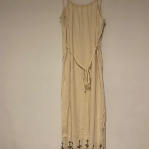 Beige långklänning med broderade detaljer - Säljer en beige långklänning med smala axelband och knyt i midjan. Klänningen har fina svarta broderade detaljer längst ner vid fållen som ger en bohemisk känsla. Perfekt för sommarens varma dagar.