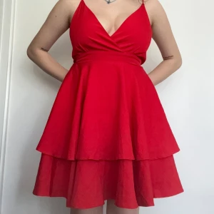 Röd volangklänning med omlott och knyt i ryggen - Säljer en röd kortklänning med omlott framtill, volangkjol och smala axelband som korsas i ryggen. Klänningen har en öppen rygg och knyt bak, perfekt för sommarens alla tillfällen.