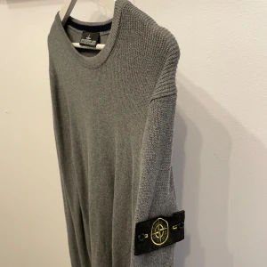 Grå stickad tröja Stone Island - Säljer en grå stickad tröja från Stone Island med klassisk patch på ärmen. Tröjan har rund halsringning och långa ärmar. Perfekt för dig som gillar stilrena och exklusiva plagg.