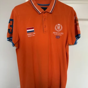 Orange pikétröja från Henri Lloyd - Säljer en orange pikétröja från Henri Lloyd med broderade detaljer och Nederländerna-flagga. Tröjan har klassisk krage med blå och vita ränder samt korta ärmar med blå kant. Perfekt för en sportig och stilren look.