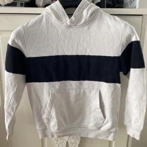 Vit och svart hoodie med bred rand barn 122/128 - Säljer en vit hoodie med en bred svart rand över bröstet och armarna. Tröjan har huva och en stor magficka framtill. Perfekt för en avslappnad stil och enkel att matcha med jeans eller joggers. Storlek står ej så jag uppskattar. Kan mäta. Det finns ngn fläck. Har ngn med fri frakt så hör av er. 