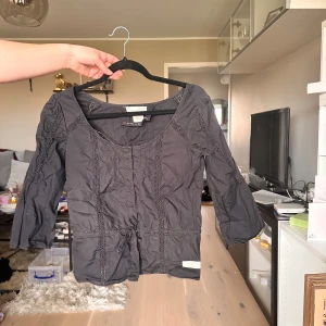 Svart blus med spetsdetaljer från Odd Molly - Svart blus från Odd Molly med trekvartsärm och dekorativa spetsdetaljer framtill och baktill. Blusen har rund halsringning och en liten rosett framtill i midjan. Perfekt för en stilren och trendig look.