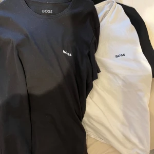 Svart och vit t-shirt från BOSS - Två stilrena t-shirts från BOSS, en svart och en vit, båda med liten BOSS-logga på bröstet. Klassisk rund hals och korta ärmar. Perfekta för en clean och enkel look.