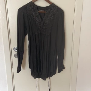 Svart spetsklänning Alice Bizous XS - Svart klänning från Alice Bizous i storlek XS med lång ärm och vacker spetsdetalj vid halsringningen. Klänningen har knappar framtill och ett lätt fallande tyg med knytband i midjan. Perfekt för dig som gillar romantiska detaljer och en bohemisk vibe.