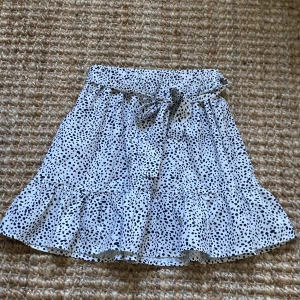 Prickig vit kjol med volang från SHEIN - Superfin kort kjol från SHEIN i vit med svarta prickar. Kjolen har en volangkant nedtill och ett sött knytband i midjan. Perfekt för dig som gillar lekfull stil och mönster. Lätt och luftig, passar perfekt till varma dagar.